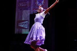 Girl in purple dress singing joyfully with her arms in the air.